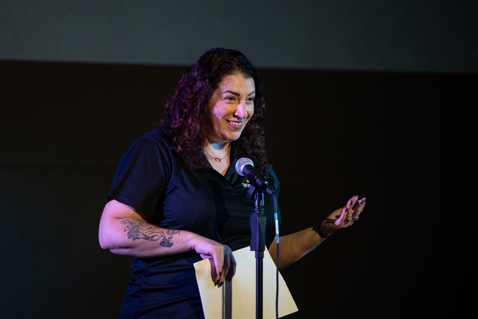 Lindsay Mastro speaking at a microphone on a dark stage